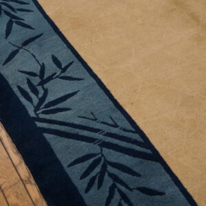 A tan rug with a navy and teal border displaying a bamboo leaf pattern rests on wood floors.