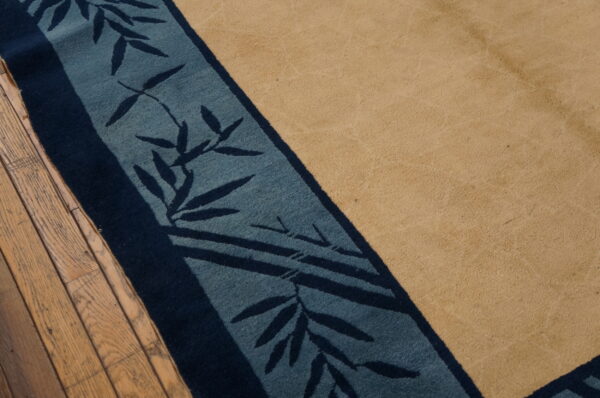 A tan rug with a navy and teal border displaying a bamboo leaf pattern rests on wood floors.