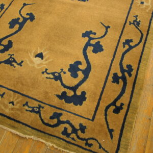 A golden-brown rug with a navy blue scrolling border rests on textured, light brown wood flooring.