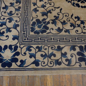 The corner of a beige pile rug features a navy floral pattern and geometric border on wooden floorboards.