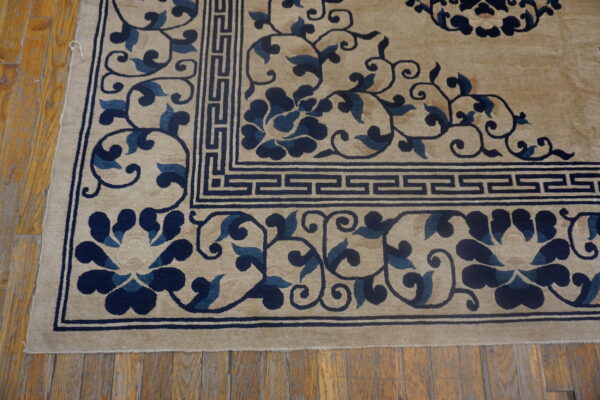 The corner of a beige pile rug features a navy floral pattern and geometric border on wooden floorboards.