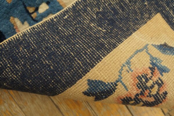 Detail of a rug showing the dark blue foundation and patterned beige pile edge over distressed wood flooring.