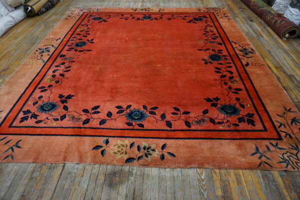 Red-orange field rug with dark blue floral patterns and a wide peach border on distressed wood floor.