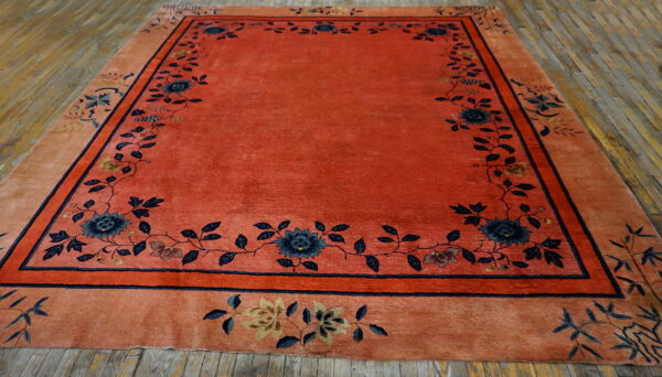 Red-orange rug with a dense navy floral border displayed on distressed wooden floorboards.