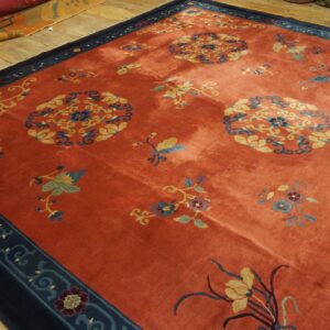 Terracotta rug with gold and blue floral designs and a navy border rests on worn wood plank flooring.