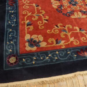 Rust-colored rug with gold floral designs and wide navy blue and teal borders rests on hardwood flooring.
