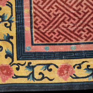 Close-up shows a rug corner with a pink geometric center, dark blue key border, and a gold field with pink and teal florals.