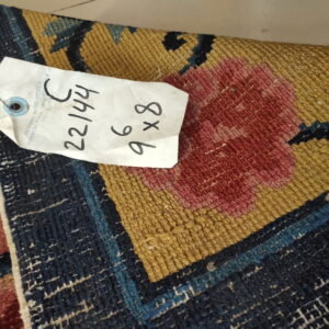 Detail of a low-pile rug displaying bold dark blue, gold, and pink woven patterns with an attached tag.