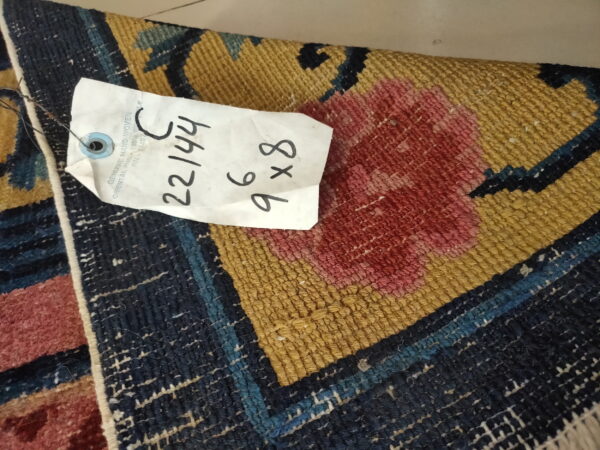 Detail of a low-pile rug displaying bold dark blue, gold, and pink woven patterns with an attached tag.