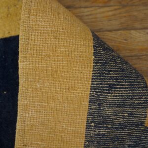 A low-pile navy and mustard rug corner is peeled back, revealing its textured woven backing on wood flooring.