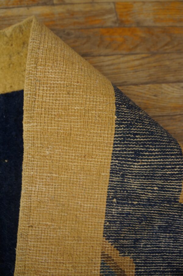 A low-pile navy and mustard rug corner is peeled back, revealing its textured woven backing on wood flooring.
