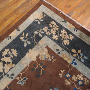 Corner of a brown floral rug with complex charcoal and tan borders on wood planks.
