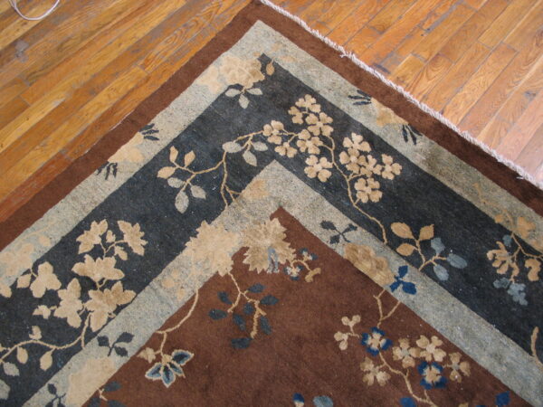 Corner of a brown floral rug with complex charcoal and tan borders on wood planks.