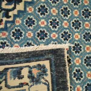 A folded rug displays a teal field with floral motifs next to a navy geometric border and cream fringe.