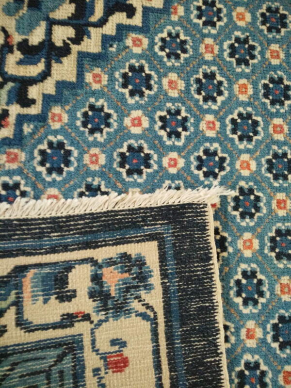 A folded rug displays a teal field with floral motifs next to a navy geometric border and cream fringe.