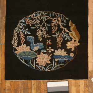 Black rug features a central round floral vignette in blue, pink, and tan on plank wood flooring.