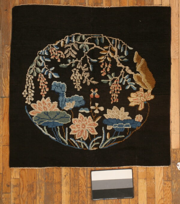 Black rug features a central round floral vignette in blue, pink, and tan on plank wood flooring.