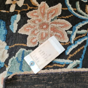 A label rests on a dark, high-pile rug with stylized flowers in peach, blue, and cream colors.
