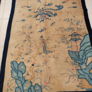 Tan rug with a navy border features an intricate scenic pattern of a bird, trees, and rocks.