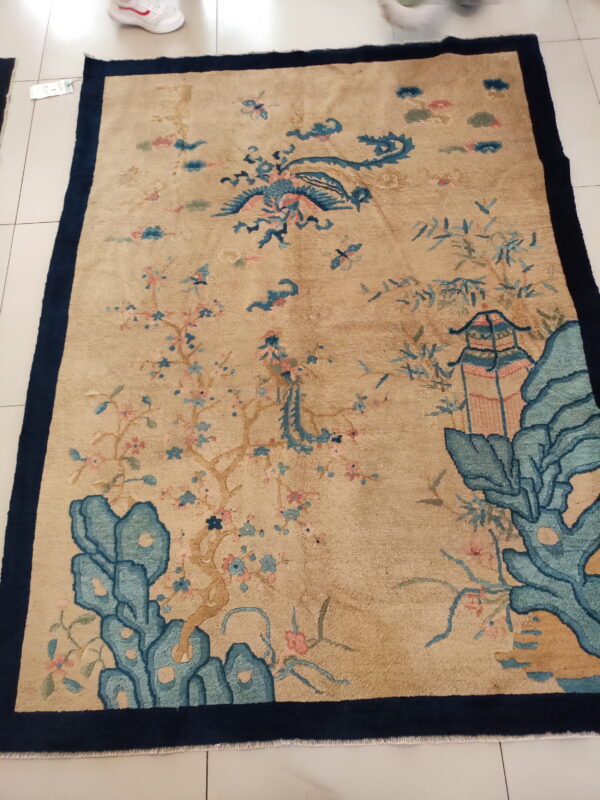 Tan rug with a navy border features an intricate scenic pattern of a bird, trees, and rocks.