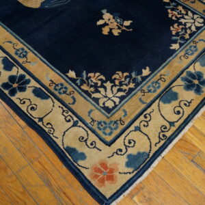 Corner view of a navy blue rug with gold and blue floral borders on distressed wood floors.
