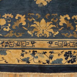 Detail of a deep blue rug showing gold floral motifs, geometric key patterns, and fringe on wood floors.
