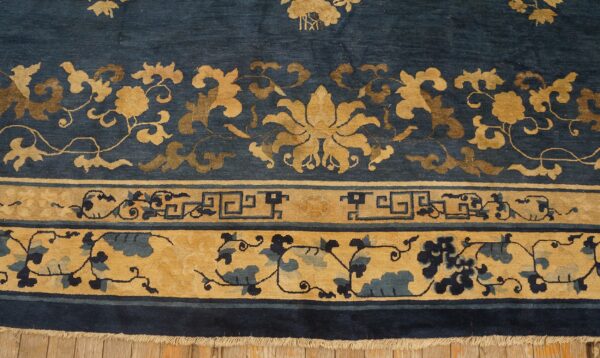 Detail of a deep blue rug showing gold floral motifs, geometric key patterns, and fringe on wood floors.