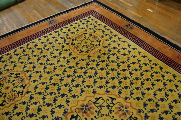Gold rug featuring repeating blue and terracotta motifs, a geometric border, and a rust-colored edge on wood flooring.