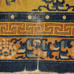 Worn, low-pile rug in yellow, navy, and terracotta with floral and geometric borders on wood planks.