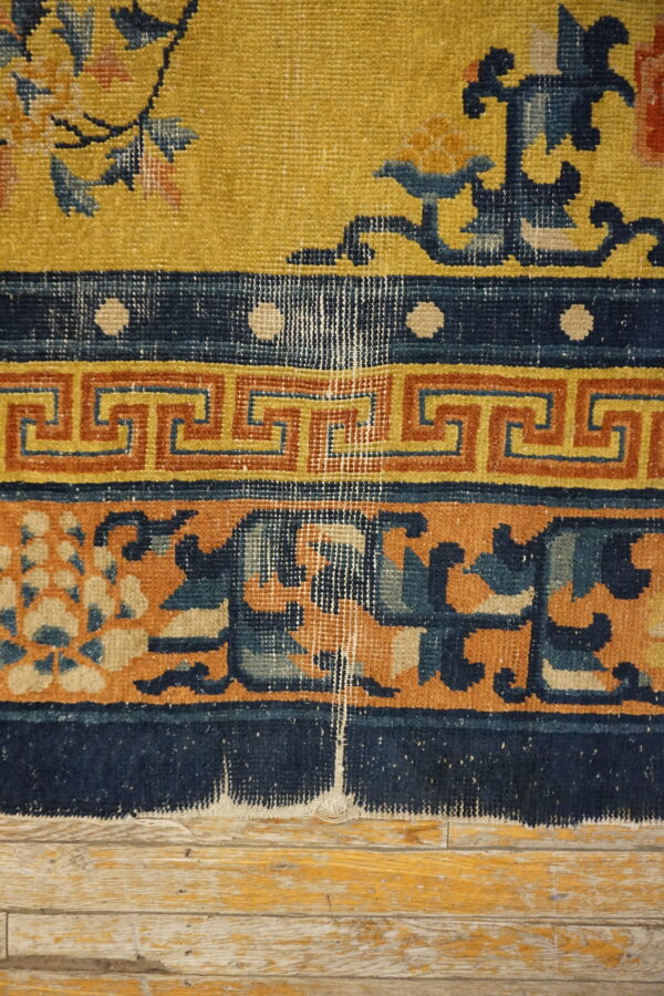 Worn, low-pile rug in yellow, navy, and terracotta with floral and geometric borders on wood planks.