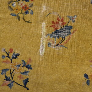 Worn gold rug displaying floral and bird patterns, a navy border, and exposed white foundation damage.
