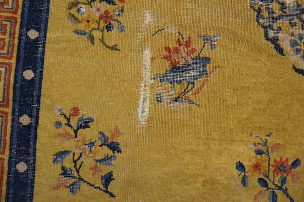 Worn gold rug displaying floral and bird patterns, a navy border, and exposed white foundation damage.