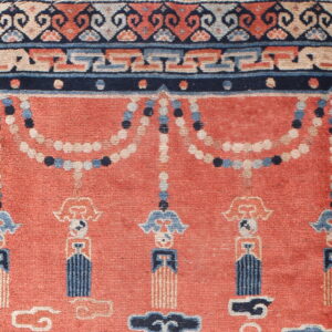 Low-pile rug detail showing a rust field with blue and beige tassels and a patterned navy border.