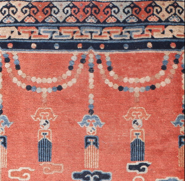 Low-pile rug detail showing a rust field with blue and beige tassels and a patterned navy border.