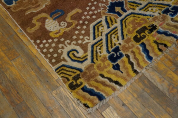 Brown rug with blue, yellow, and cream geometric and scroll patterns on worn wood floorboards.