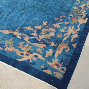 Teal rug with a detailed landscape border pattern in tan, rust, and navy blue on a concrete floor.