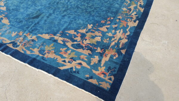 Teal rug with a detailed landscape border pattern in tan, rust, and navy blue on a concrete floor.
