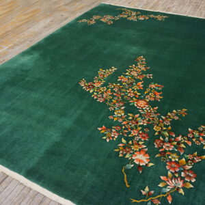 Green rug with plush pile displaying sweeping orange and yellow floral designs on worn hardwood.