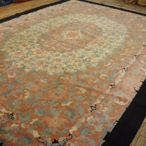 Large low-pile rug featuring faded coral and cream ornate patterns, bordered by solid black on wood flooring.