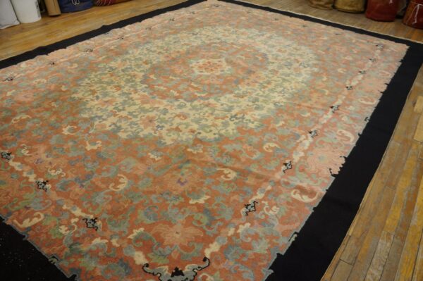 Large low-pile rug featuring faded coral and cream ornate patterns, bordered by solid black on wood flooring.