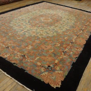 Rust and cream floral rug featuring a black border and central medallion, displayed on worn wooden plank flooring.