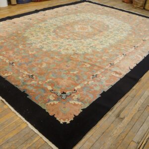 Muted coral and cream patterned rug with a wide black border displayed on distressed wood flooring.