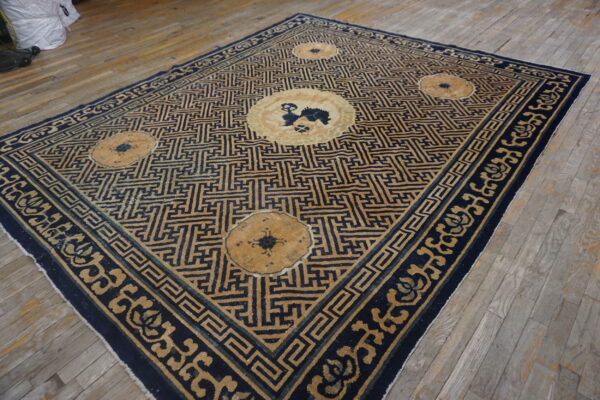 Rectangular low-pile rug featuring a blue and tan meander pattern with medallions on light wood flooring.