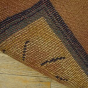 A tightly woven rug corner is lifted, showing russet and gold striped patterns over light wood flooring.
