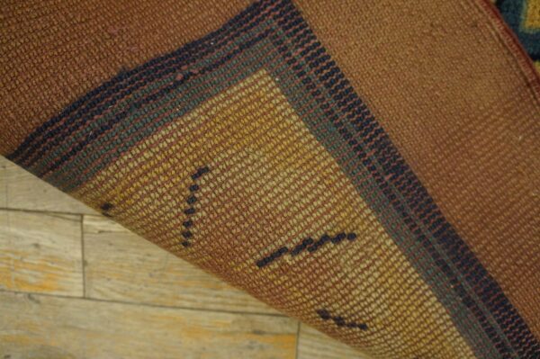 A tightly woven rug corner is lifted, showing russet and gold striped patterns over light wood flooring.