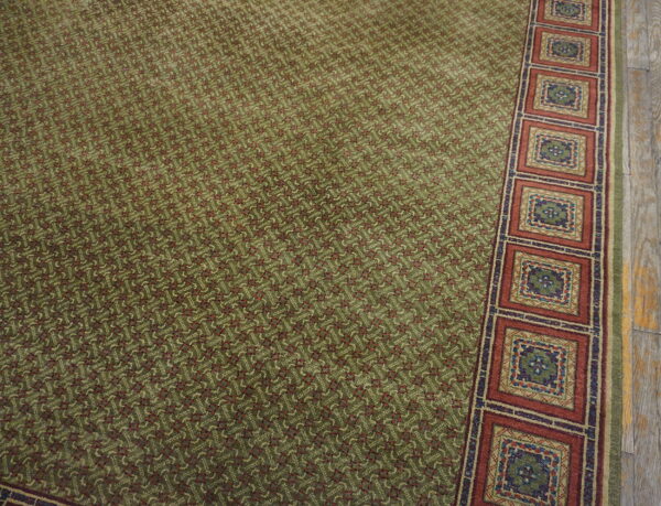 Detail of an olive green rug showing a geometric field and a border of rust-colored square medallions on wood flooring.