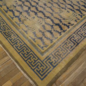 Close-up of a distressed gold and blue floral rug with a bold Greek key border on rustic wood flooring.