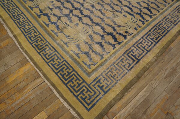 Close-up of a distressed gold and blue floral rug with a bold greek key border on rustic wood flooring.