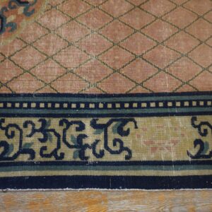 Close-up of a low-pile rug featuring a salmon diamond lattice field and multi-band border with navy scrollwork on wood flooring.