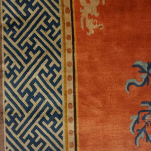 Rust rug with blue botanical patterns and tan geometric key borders resting on light wood flooring.